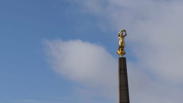 A golden statue of a woman holding a wreath atop a tall column against a blue sky with clouds.