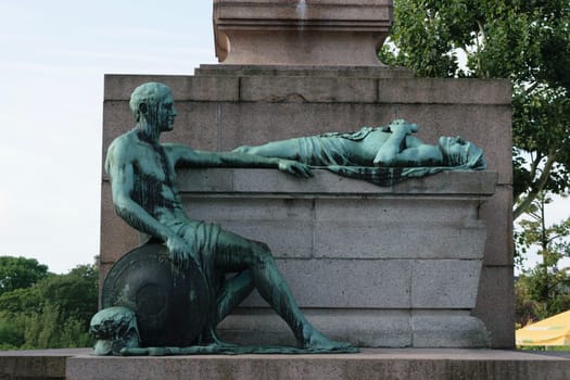 A bronze statue depicting a seated man holding a shield and a lying figure on a stone pedestal in an outdoor setting.