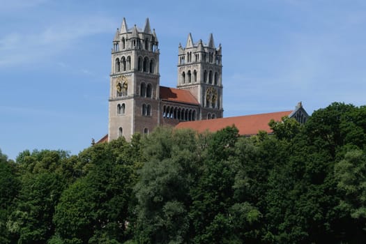 A historic church with twin towers and a red roof, partially obscured by lush green trees, under a clear blue sky.