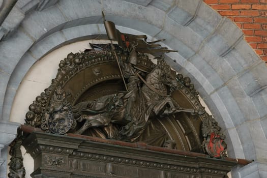 A detailed bronze relief sculpture depicting a knight on horseback holding a flag, set within an arched stone frame. The sculpture includes intricate details and heraldic symbols.