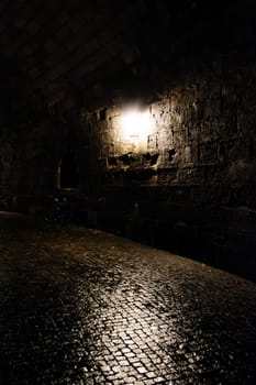 A dimly lit cobblestone street under a stone archway at night, with a single wall-mounted lamp illuminating part of the scene.