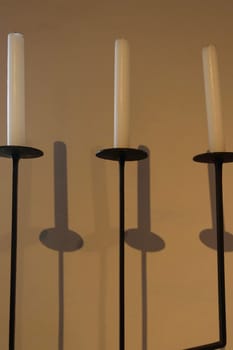 Three unlit white candles on black metal candle holders casting shadows on a beige wall.