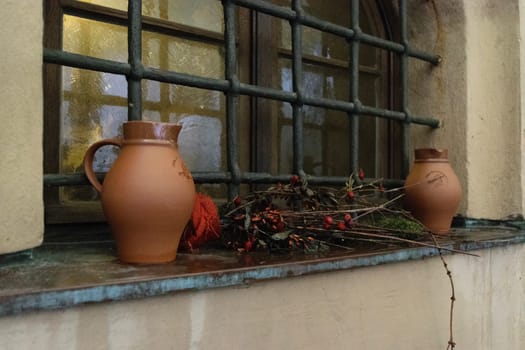 Two ceramic jugs and a bundle of dried branches with berries placed on a windowsill with metal bars. The window has an arched top and is set in a rustic, weathered wall.