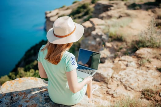 A woman is sitting on a rock with a laptop open in front of her. She is wearing a straw hat and a green dress. The scene suggests a relaxed and leisurely atmosphere