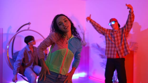 Attractive happy dancer looking at camera while smart woman moving at rhyme with neon light. Professional hispanic performer break dancing while wearing colorful cloth with diverse friend. Regalement.