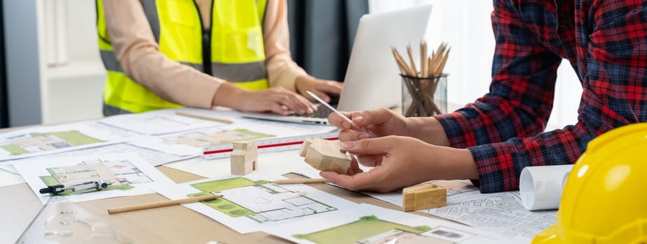 Professional architect and engineer collaborate to design eco-friendly house on meeting table with blueprint, wooden house block and tree model scatter around. Closeup. Delineation.