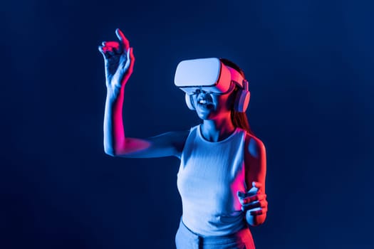 Smart female standing in cyberpunk neon light wear VR headset connecting metaverse, futuristic cyberspace community technology. Elegant woman use finger point generated virtual object. Hallucination.