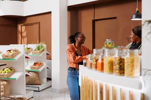 Black female cashier serves a client at an eco-friendly grocery store, offering freshly harvested, sustainable food. African american customer speaking with vendor at checkout counter.