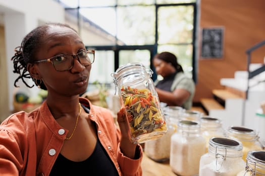 African american female influencer vlogging in bio food store while holding glass container of pasta. POV of black woman describing fresh product in a jar towards camera for social media.