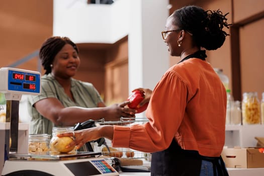 Young woman at checkout in zero waste shop, buying locally grown ecofriendly vegetables from friendly storekeeper. Female customer purchasing organic chemicals free groceries.