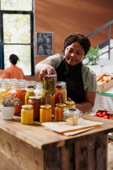 Female storekeeper selling jars of food, including pickles, in an environmentally conscious grocery shop. African american seller organizing reusable containers with organic preserved products.