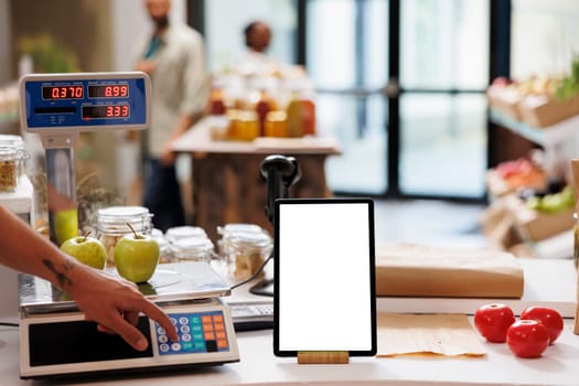 Digital tablet displaying a white screen while salesperson is using an electronic weight scale nearby. Smart device with isolated chromakey template is vertically positioned on checkout counter.