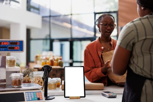 A lady wearing glasses converses with a vendor at the payment counter, while a digital device showcases a blank white screen featuring eco-friendly store advertisements. The tablet displays an empty chromakey mockup template