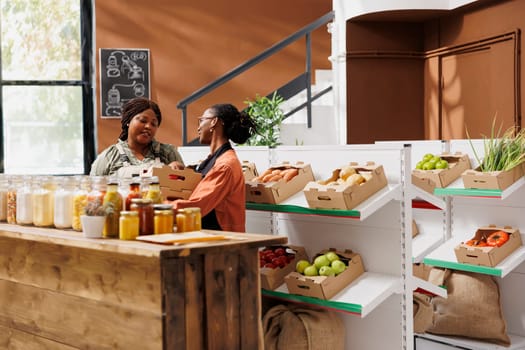 African American vendor sells fresh, organic fruits and vegetables at green grocery store. Local farmer giving crates of freshly harvested organic produce to female shopkeeper.