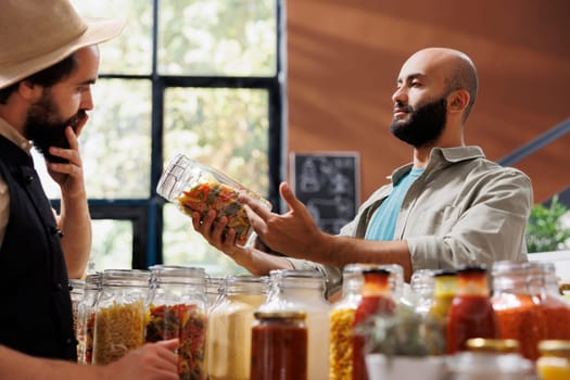 A young man explores an eco-friendly store, looking at a variety of fresh organic pantry products in reusable packaging. A Middle Eastern client examines jars of pasta and grains.