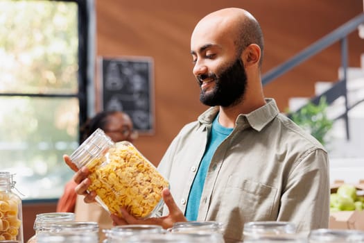 Client buys organic sustainable food in ecological local neighborhood store. Selective focus on middle eastern man looking at glass jar filled with natural organic cereal.