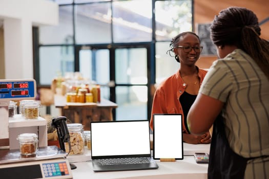 Customer at checkout counter with fresh, organic food products. Vendor promotes eco friendly, zero waste goods using laptop and tablet, white screen provides blank template for digital advertisement.