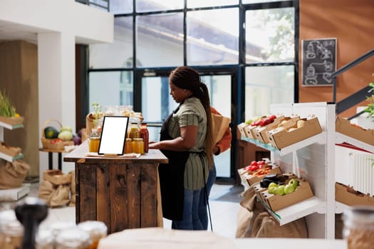 Vendor stands by table filled with glass jars filled with organic products while interacting with someone in environmentally friendly grocery store. Digital tablet on desk displaying white screen.