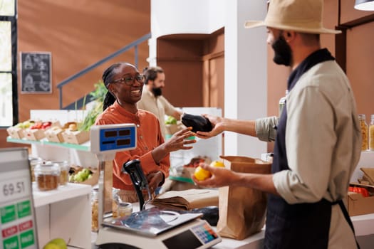 Smiling african american female customer handing over locally grown eggplant to middle eastern cashier for weighing. Black woman being assisted by male vendor at checkout counter.