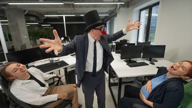 An elderly Caucasian man in a clown costume amuses two Caucasian women in the office