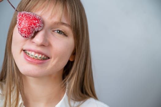 Cute woman with braces on her teeth holds a candy in the form of a heart on white background.