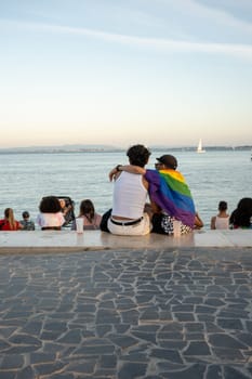 Back view of gay couple sitting with arms around each other on promenade near sea under sky. LGBTQ individuals spending leisure time together, with rainbow flag showing support for LGBT community.
