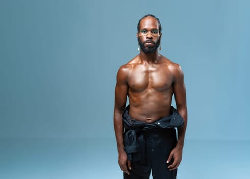 Portrait of shirtless African American gay man with light makeup standing isolated on blue background. Muscular black LGBTQ person in trendy pants looking at camera.