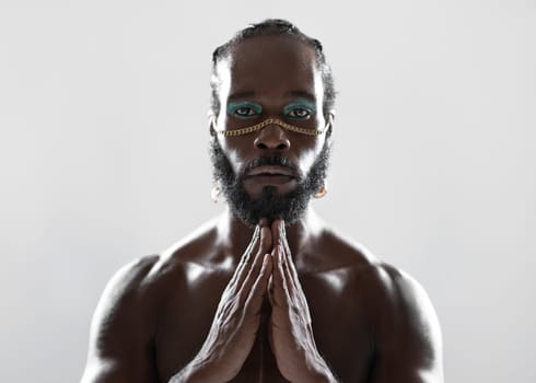 Confident African-American bearded gay man with bright makeup joining hands, isolated on white background. Close-up portrait of gay African man making namaste gesture and looking at camera.