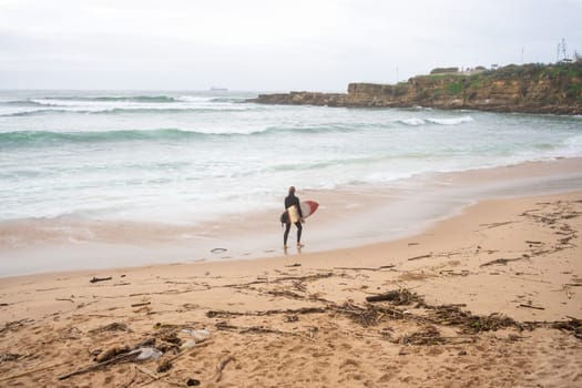 Anonymous surfer carrying surfboard gazing at sea waves rushing towards the shore. Unknown person is ready for surfing session while standing on sandy beach, engaging in extreme water sport.