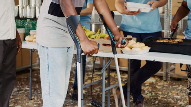 African American woman volunteer assists and supports crippled man at outdoor food bank. The hungry, poor and homeless people receive hunger relief assistance from multiethnic voluntary individuals.