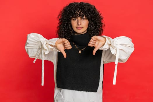 Dislike. Upset unhappy Caucasian woman showing thumbs down sign gesture, expressing discontent, disapproval, dissatisfied negative feedback. Pretty beautiful girl indoors isolated on red background