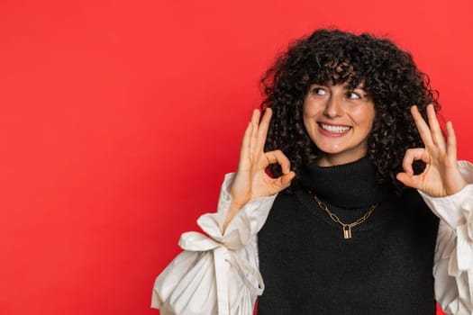 Okay. Happy Caucasian young woman with curly hair looking approvingly at camera showing ok gesture, like sign positive feedback, approve something good, celebrate victory. Girl on red background