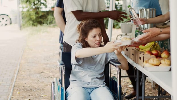 Caucasian wheelchair-bound woman receiving free food from charity workers at outdoor non-profit program. Hunger relief volunteer team handing out provisions to needy homeless and disabled people.