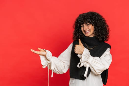 Caucasian beautiful woman showing thumbs up and pointing left empty place advertising area for commercial text copy space for goods promotion advertisement. Girl isolated on red background. Lifestyle