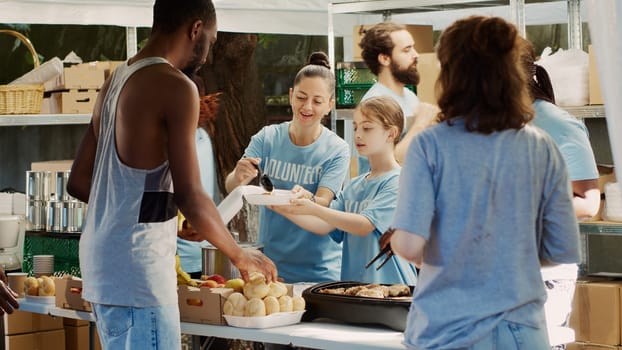 Young girl assisted by fellow volunteers, packing warm free food for handing out to underprivileged. Multiethnic charity workers at homeless shelter freely give away meals to poor and spread joy.