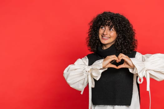 Woman in love. Smiling attractive Caucasian young woman with curly hair makes heart gesture demonstrates love sign expresses good positive feelings and sympathy. Girl isolated on red background