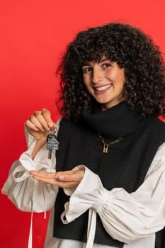 Young woman with curly hair real estate agent lifting hand showing the keys of new home house apartment, buying or renting property, mortgage loan, insurance. Girl isolated on red background. Vertical