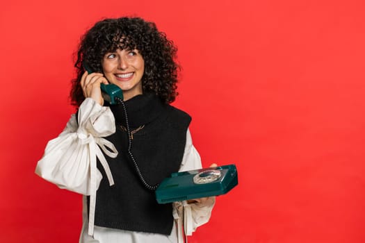 Hey you, call me. Caucasian young woman with curly hair talking on wired landline vintage telephone of 80s, advertising proposition of conversation, online shopping. Girl isolated on red background