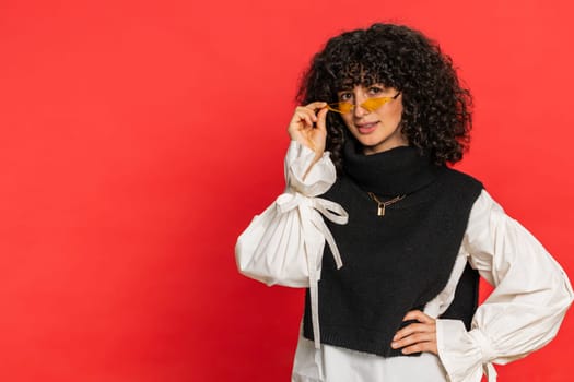 Stylish smiling Caucasian young woman with curly hair wearing orange sunglasses glad expression looking at camera dreaming, resting relaxation feel satisfied good news. Girl isolated on red background