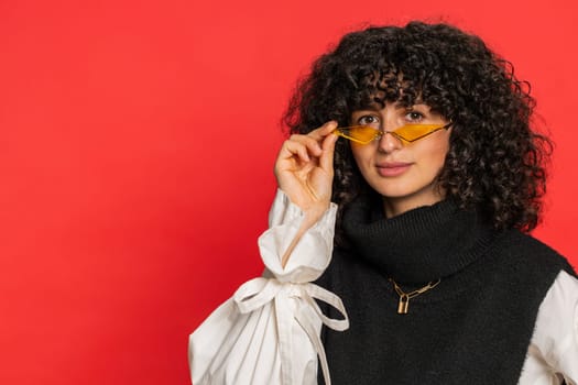 Stylish smiling Caucasian young woman with curly hair wearing orange sunglasses glad expression looking at camera dreaming, resting relaxation feel satisfied good news. Girl isolated on red background