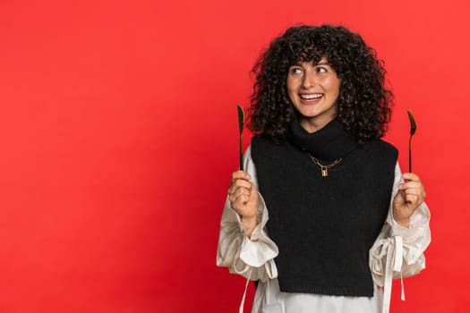 Ready to eat. Hungry Caucasian woman with curly hair waiting for serving dinner dishes with with restlessness holding cutlery fork knife, will appreciate delicious restaurant meal on red background