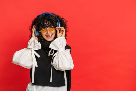 Caucasian young woman with curly hair listening music on headphones, dancing disco fooling around having fun expressive gesticulating hands. Music app advertisement. Girl isolated on red background