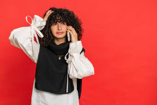 Disinterested woman with curly hair having annoyed boring talk on smartphone. Girl tired sleepy freelancer talking on mobile phone with friend, making online conversation isolated on red background