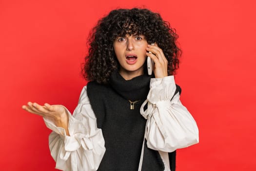 Disinterested woman with curly hair having annoyed boring talk on smartphone. Girl tired sleepy freelancer talking on mobile phone with friend, making online conversation isolated on red background