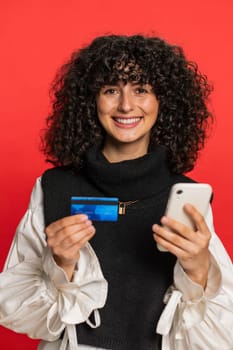 Happy Caucasian young woman with curly hair using credit bank card and smartphone while transferring spending money, purchases online shopping, ordering food delivery. Girl isolated on red background