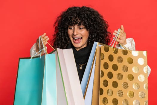 Happy Caucasian young woman with curly hair showing shopping bags, advertising discounts, smiling looking amazed with low prices, purchase on Black Friday holidays. Girl isolated on red background