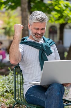 Handsome mature man using laptop computer celebrating win good message news, lottery jackpot victory, giveaway online outdoors. Happy bearded guy tourist sits in city cafe terrace. Town lifestyles.