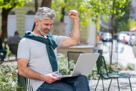 Handsome mature man using laptop computer celebrating win good message news, lottery jackpot victory, giveaway online outdoors. Happy bearded guy tourist sits in city cafe terrace. Town lifestyles.