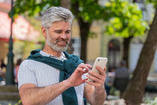 Happy smiling mature man using smartphone typing text messages tired looking up thinking and browsing internet social media web app working chatting online outdoor. Joyful guy in urban city street