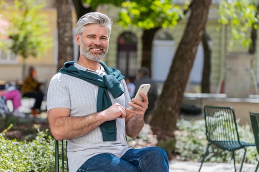 Handsome mature man using smartphone typing text messages tired looking up thinking and browsing internet social media web app working chatting online outdoor. Guy in urban city street. Lifestyles.